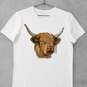 May include: White t-shirt with a brown and white Highland cow illustration. The cow has long, shaggy hair and is looking directly at the viewer.