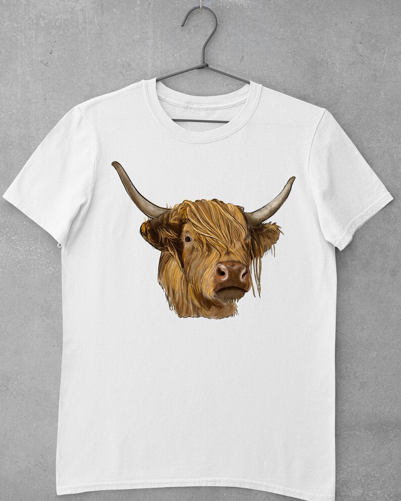 May include: White t-shirt with a brown and white Highland cow illustration. The cow has long, shaggy hair and is looking directly at the viewer.