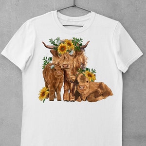 May include: A white t-shirt with a brown Highland cow and two calves. The cow is wearing a sunflower crown. The calves are also wearing sunflower crowns. The design is in a cartoon style.