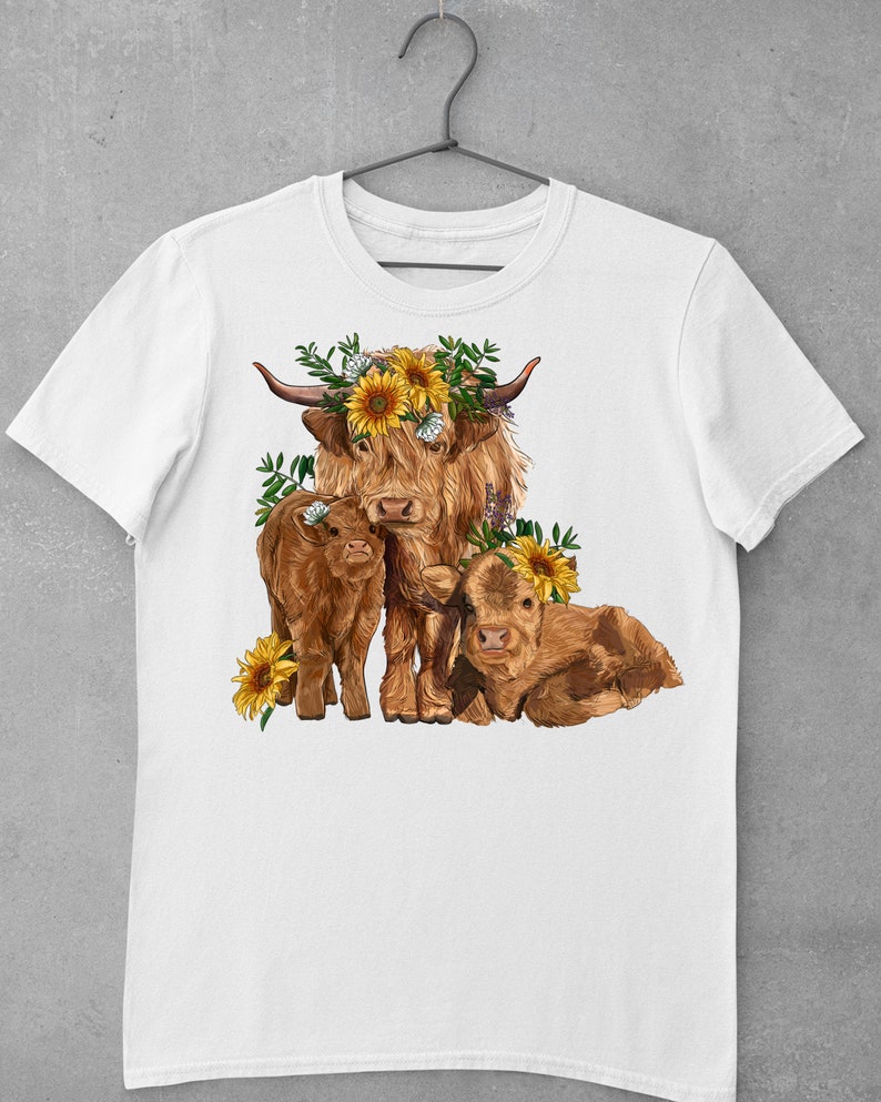 May include: A white t-shirt with a brown Highland cow and two calves. The cow is wearing a sunflower crown. The calves are also wearing sunflower crowns. The design is in a cartoon style.