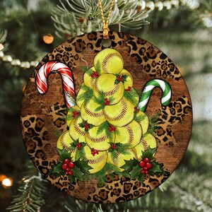 Pode incluir: Um enfeite de Natal redondo com fundo de estampa de leopardo. Uma árvore de Natal é feita de bolas de softball amarelas e brancas com azevinho verde e bagas vermelhas. Dois bastões de caramelo estão de cada lado da árvore, um vermelho e branco e o outro verde e branco.