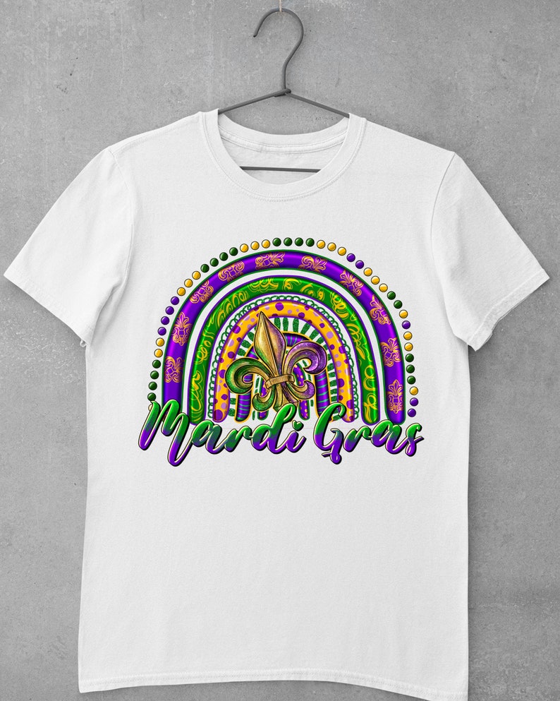 Puede incluir: Camiseta blanca con un dise&ntilde;o de arco&iacute;ris de Mardi Gras colorido con una flor de lis y el texto "Mardi Gras".