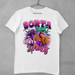 May include: White t-shirt featuring a graphic design of a woman's legs wearing purple shoes and gold jewellery. The text "Sorta Sweet Sorta Spooky" is written in pink and purple letters.