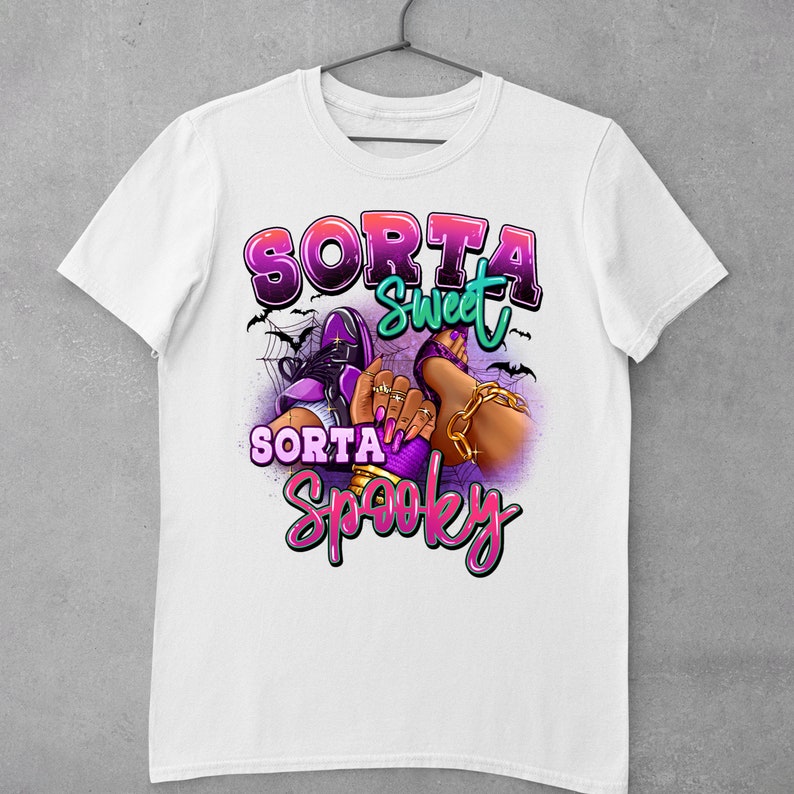 May include: White t-shirt featuring a graphic design of a woman's legs wearing purple shoes and gold jewellery. The text "Sorta Sweet Sorta Spooky" is written in pink and purple letters.