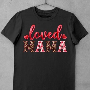 K&ouml;nnte beinhalten: Schwarzes T-Shirt mit dem Text "loved" in Rot mit einem Herz auf jeder Seite und dem Wort "MAMA" in gro&szlig;en Buchstaben mit einem Leopardenmuster und rosa Herzen.