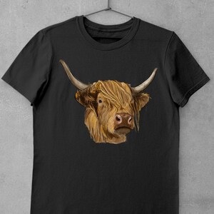 May include: A black t-shirt with a brown and white Highland cow illustration. The cow has long, shaggy hair and is looking directly at the viewer.