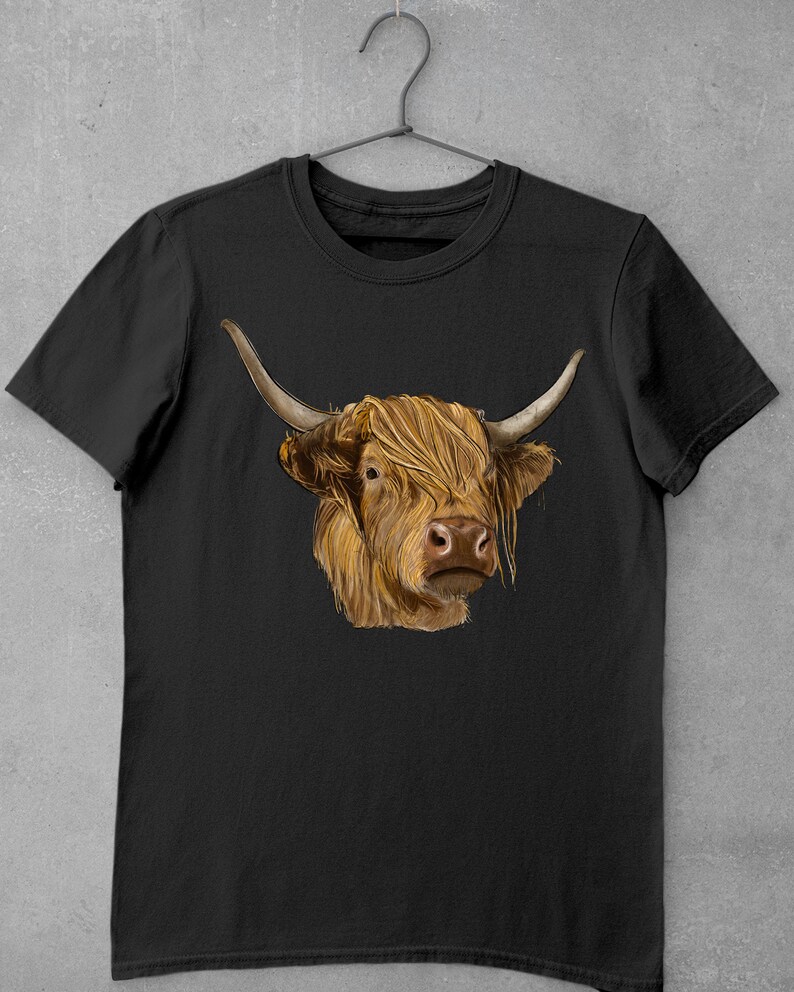 May include: A black t-shirt with a brown and white Highland cow illustration. The cow has long, shaggy hair and is looking directly at the viewer.