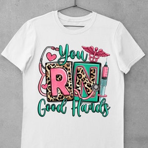 K&ouml;nnte beinhalten: Wei&szlig;es T-Shirt mit einem gr&uuml;nen, rosa und schwarzen Grafikdesign, auf dem "You're RN Good Hands" steht. Die Grafik enth&auml;lt ein Stethoskop, eine Spritze und einen &Auml;skulapstab.