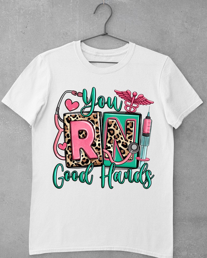K&ouml;nnte beinhalten: Wei&szlig;es T-Shirt mit einem gr&uuml;nen, rosa und schwarzen Grafikdesign, auf dem "You're RN Good Hands" steht. Die Grafik enth&auml;lt ein Stethoskop, eine Spritze und einen &Auml;skulapstab.