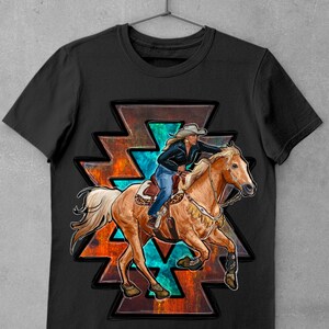 May include: Black t-shirt featuring a graphic of a woman riding a horse in a rodeo. The design includes a Native American-inspired geometric shape with turquoise and orange accents. The rider wears a cowboy hat and blue jeans.