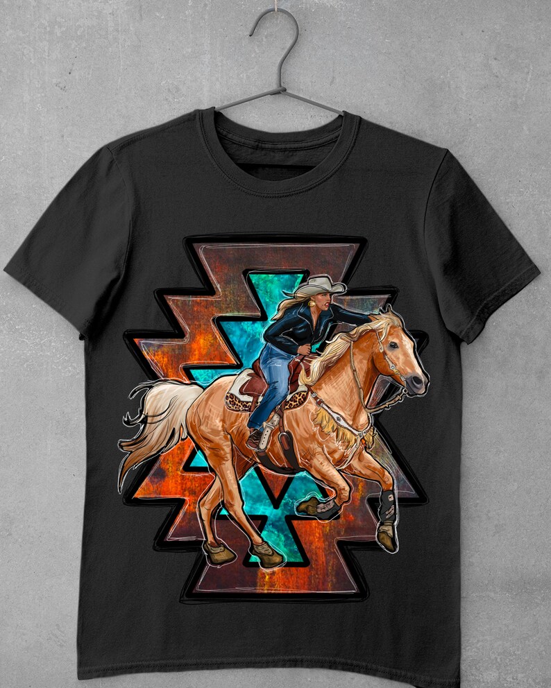 May include: Black t-shirt featuring a graphic of a woman riding a horse in a rodeo. The design includes a Native American-inspired geometric shape with turquoise and orange accents. The rider wears a cowboy hat and blue jeans.