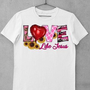 May include: White t-shirt featuring the word "LOVE" in a decorative font with a red heart balloon and sunflowers. The word "LOVE" has a pink leopard print, pink plaid, and black and pink cow print design. The phrase "Like Jesus" is written in pink script.
