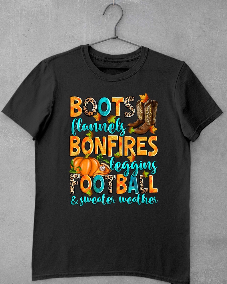May include: Black t-shirt with a graphic design featuring the text "BOOTS, flannels, BONFIRES, leggings, FOOTBALL & sweater weather" in turquoise and orange with autumn-themed imagery.