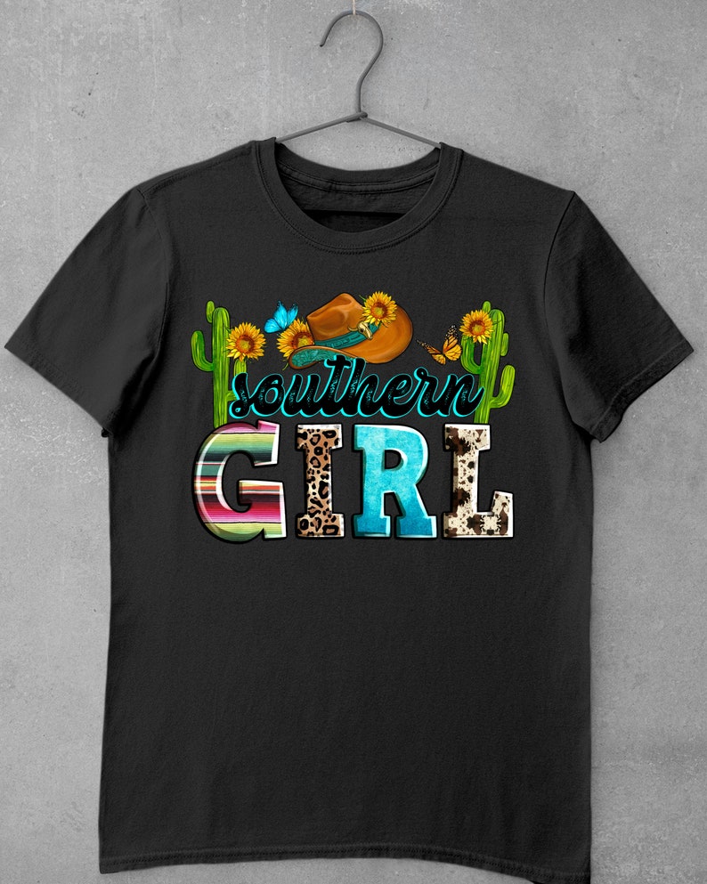 Pode incluir: Camiseta preta com um design gr&aacute;fico de um chap&eacute;u de cowboy, girass&oacute;is, borboletas e cactos. O texto "Southern Girl" est&aacute; escrito em letras coloridas com diferentes padr&otilde;es.