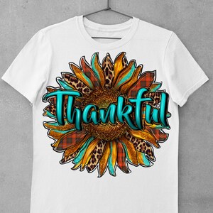 May include: White t-shirt with a turquoise, orange, and brown sunflower design. The sunflower has a leopard print and plaid pattern. The word "Thankful" is written in turquoise letters.