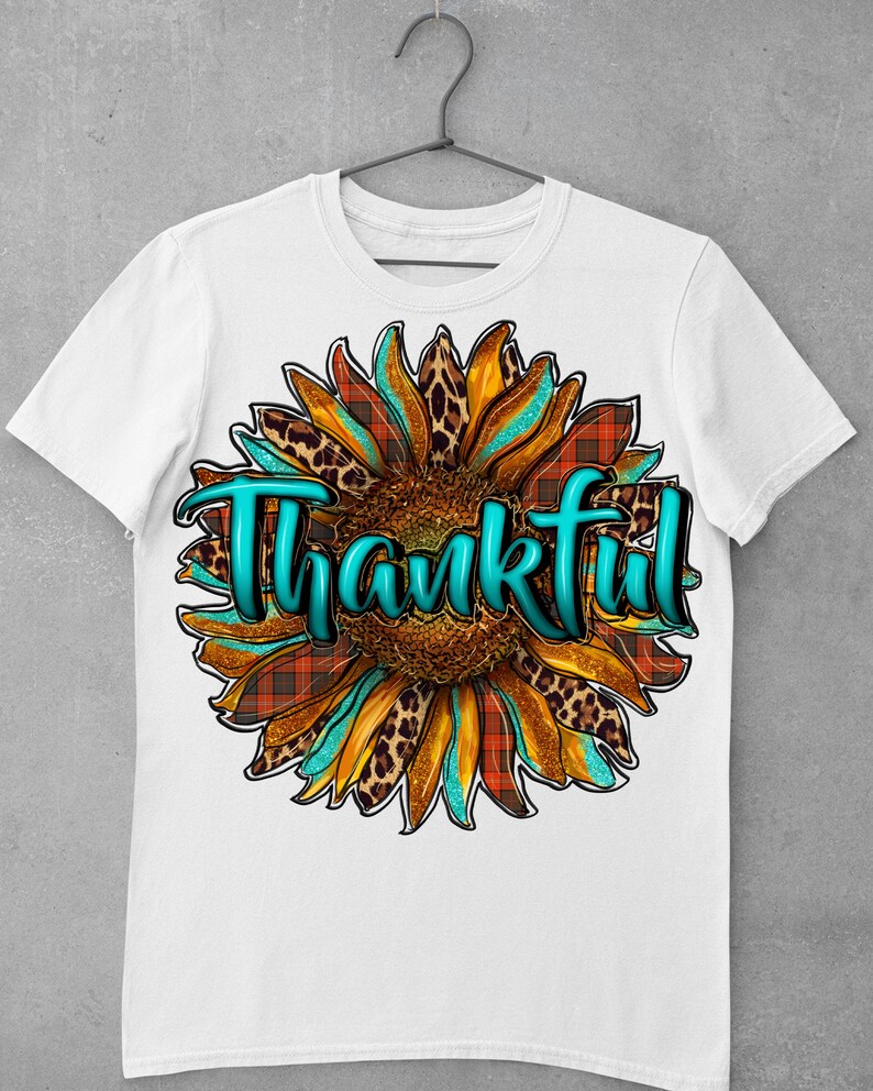 May include: White t-shirt with a turquoise, orange, and brown sunflower design. The sunflower has a leopard print and plaid pattern. The word "Thankful" is written in turquoise letters.