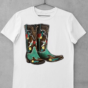 K&ouml;nnte beinhalten: Wei&szlig;es T-Shirt mit einem Aufdruck von einem Paar brauner Cowboystiefel mit bunter Stickerei. Die Stiefel haben einen gr&uuml;nen Schaft und eine spitze Zehenpartie.