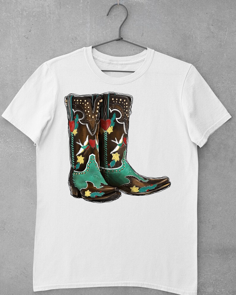 K&ouml;nnte beinhalten: Wei&szlig;es T-Shirt mit einem Aufdruck von einem Paar brauner Cowboystiefel mit bunter Stickerei. Die Stiefel haben einen gr&uuml;nen Schaft und eine spitze Zehenpartie.