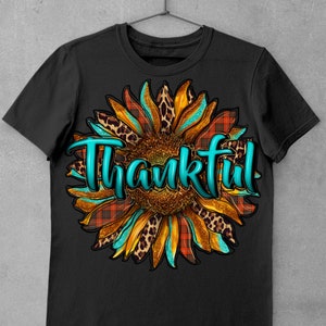 May include: Black t-shirt with a turquoise "Thankful" text overlayed on a sunflower graphic with orange, brown, and black leopard print and red and black plaid patterns.