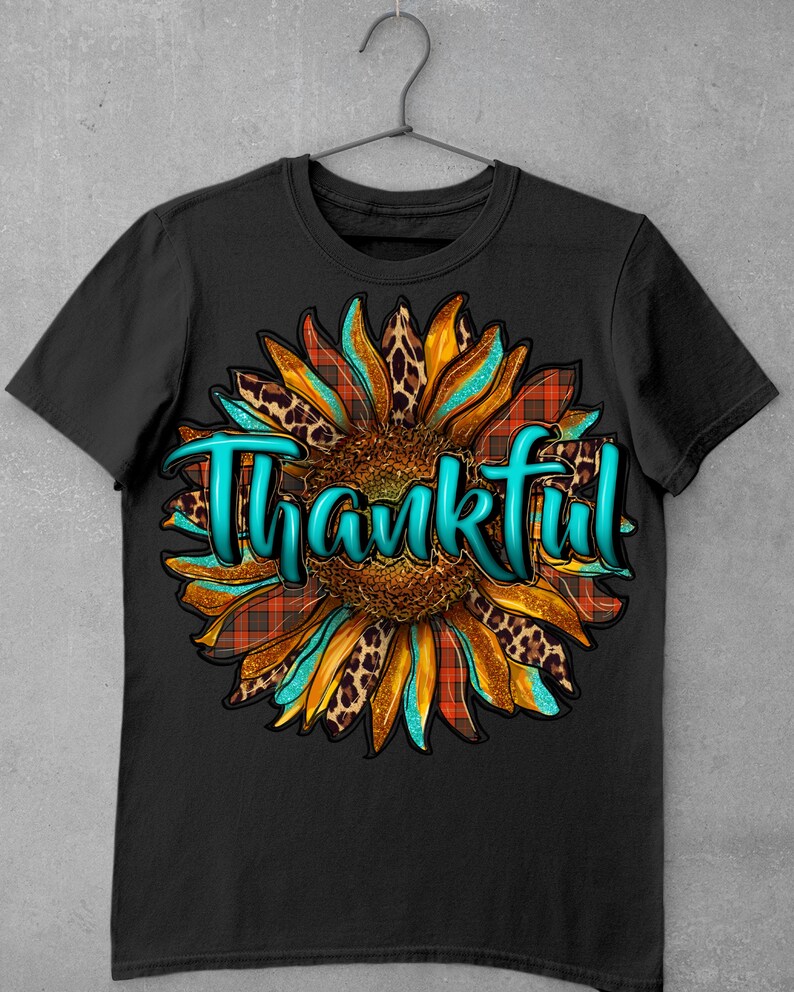 May include: Black t-shirt with a turquoise "Thankful" text overlayed on a sunflower graphic with orange, brown, and black leopard print and red and black plaid patterns.