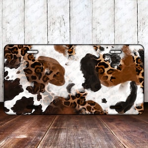 May include: Rectangular license plate featuring a cowhide and leopard print design. The pattern includes patches of brown, black, and white, with leopard spots in shades of brown and tan. The plate has a glossy finish.