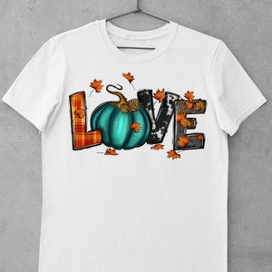 May include: White t-shirt with a graphic of the word "LOVE" made of different patterned fabrics. The "O" is a teal pumpkin with a brown stem and a pine cone on top. There are orange leaves scattered around the letters.