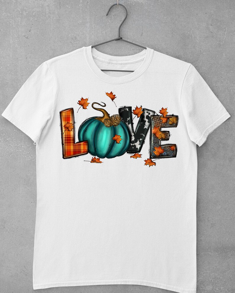 May include: White t-shirt with a graphic of the word "LOVE" made of different patterned fabrics. The "O" is a teal pumpkin with a brown stem and a pine cone on top. There are orange leaves scattered around the letters.