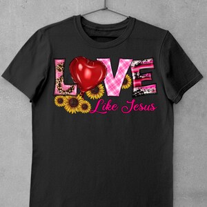 May include: Black t-shirt with the words "LOVE Like Jesus" in a decorative font. The word "LOVE" features a red heart, leopard print, and pink plaid patterns. Yellow sunflowers are at the bottom.