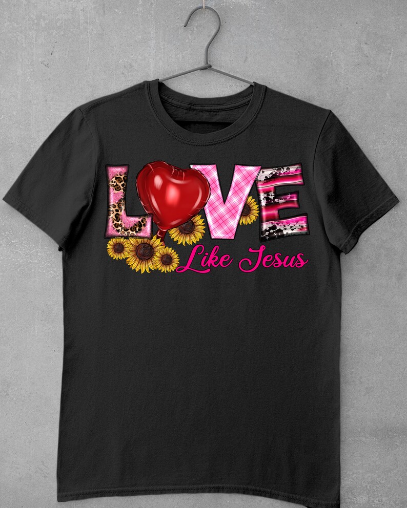 May include: Black t-shirt with the words "LOVE Like Jesus" in a decorative font. The word "LOVE" features a red heart, leopard print, and pink plaid patterns. Yellow sunflowers are at the bottom.