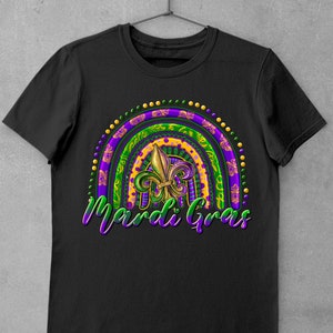 Puede incluir: Camiseta negra con un dise&ntilde;o de arco&iacute;ris de Mardi Gras morado, verde y dorado con una flor de lis en el centro. El texto "Mardi Gras" est&aacute; escrito debajo del arco&iacute;ris.