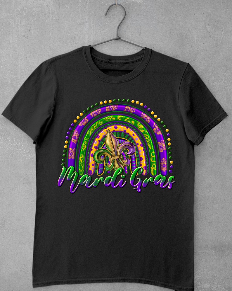 Puede incluir: Camiseta negra con un dise&ntilde;o de arco&iacute;ris de Mardi Gras morado, verde y dorado con una flor de lis en el centro. El texto "Mardi Gras" est&aacute; escrito debajo del arco&iacute;ris.