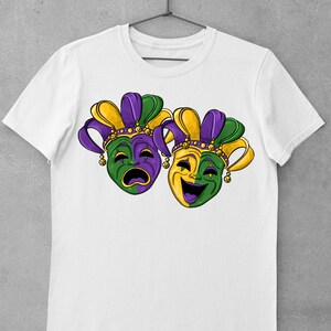May include: White t-shirt with a purple, green, and gold design of two Mardi Gras masks. One mask is crying and the other is laughing.