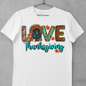 May include: White t-shirt with the word "LOVE" spelled out in colorful patterned letters. The letter "O" features a Thanksgiving turkey. The text "Thanksgiving" is written in turquoise below the word "LOVE".