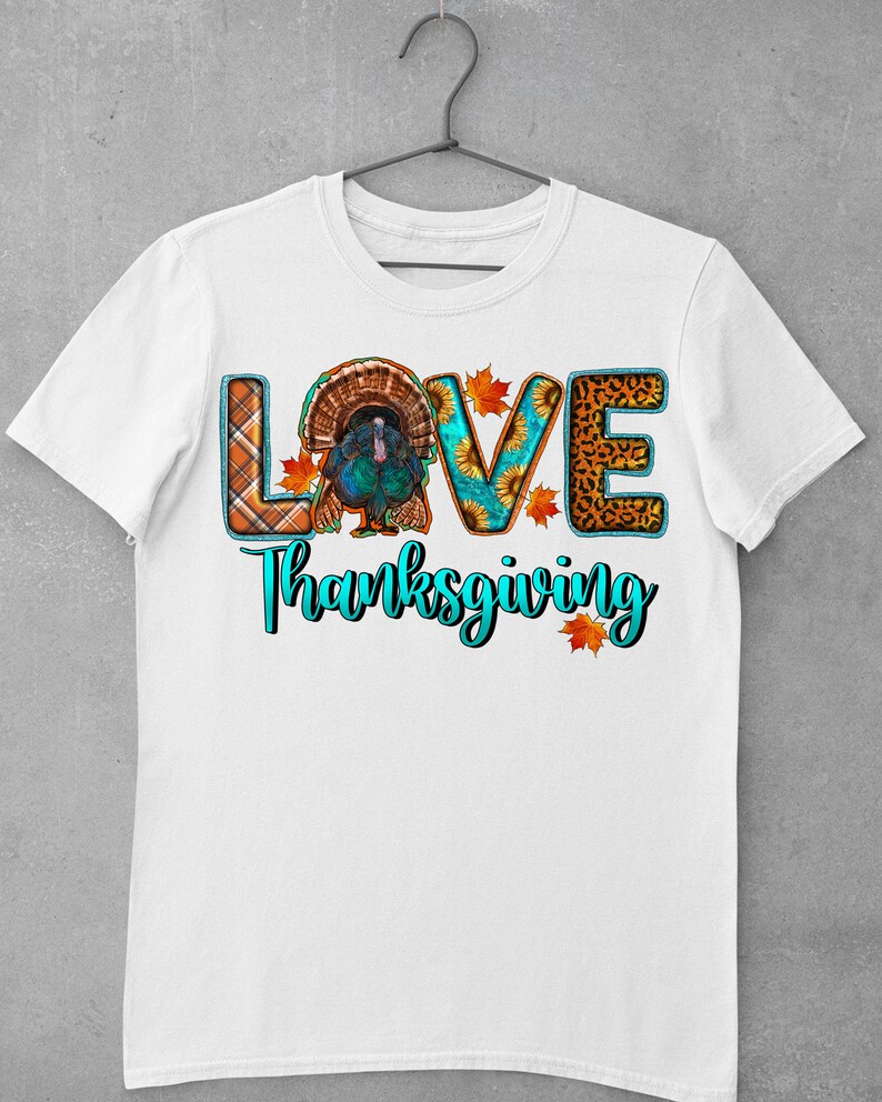 May include: White t-shirt with the word "LOVE" spelled out in colorful patterned letters. The letter "O" features a Thanksgiving turkey. The text "Thanksgiving" is written in turquoise below the word "LOVE".