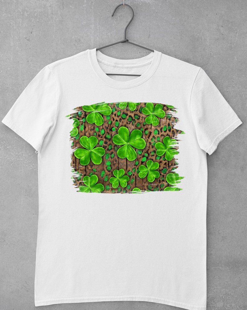 May include: White t-shirt with a green shamrock and leopard print design on a wood background.