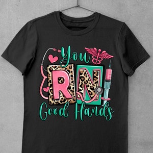 K&ouml;nnte beinhalten: Schwarzes T-Shirt mit einer gr&uuml;nen und pinkfarbenen Grafik, auf der "You RN! Good Hands" steht. Die Grafik enth&auml;lt ein Stethoskop, eine Spritze und einen &Auml;skulapstab.