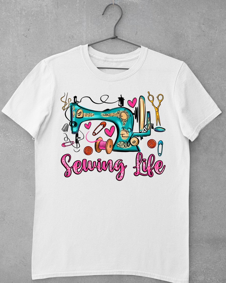 Op de afbeelding: Wit T-shirt met een turquoise naaimachine-afbeelding en de tekst "Sewing Life" in roze glitterletters. De afbeelding bevat roze harten, een schaar, knopen en een spoel met garen.