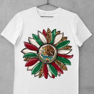 May include: White t-shirt with a green, white, and red flower design. The center of the flower features the Mexican coat of arms with an eagle perched on a cactus. The flower petals are accented with a leopard print.