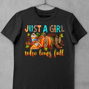 May include: Black t-shirt with the text "Just a girl who loves fall" in white and blue. The text is surrounded by fall-themed imagery, including a pumpkin, pie, boots, and a cup of coffee.