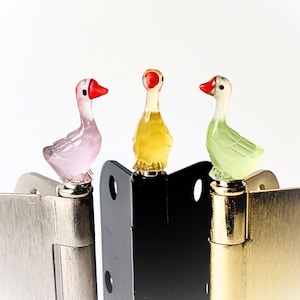 May include: Three decorative goose-shaped cabinet hinge toppers in pink, yellow, and green. Each goose has a red beak and is mounted on a metal hinge. The hinges are in silver, black, and gold.