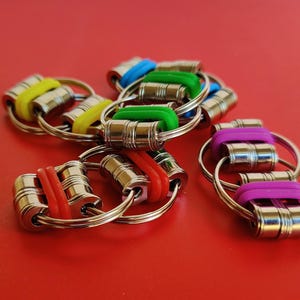 May include: A set of six colorful keychains with metal rings and colorful rubber bands. The keychains are made of metal and have a cylindrical shape. The rubber bands are in red, green, yellow, blue, and purple.