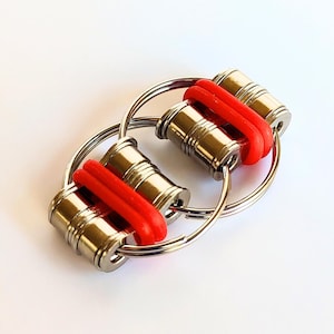 May include: A silver fidget toy with two rotating cylinders connected by three interlocking rings. The cylinders have red rubber bands around them.
