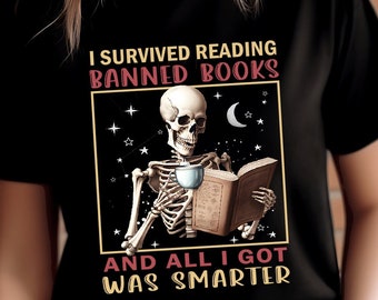 May include: A black t-shirt with a graphic of a skeleton reading a book and holding a cup. The text on the shirt reads "I survived reading banned books and all I got was smarter."