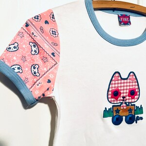 May include: White t-shirt with a blue and pink trim. The sleeves are pink with a bandana print. The front of the shirt features an applique of a cartoon cat wearing a pink and white gingham dress and pulling a wooden wagon. The wagon has two wheels and a blue star on the side. The text "FINE" is on a red tag attached to the shirt.