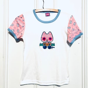 May include: White t-shirt with a blue and pink patterned trim on the sleeves. The shirt features a cartoon cat wearing a red and white checked dress and holding a box with stars. The text "Fine" is on the tag and on the bottom of the cat.