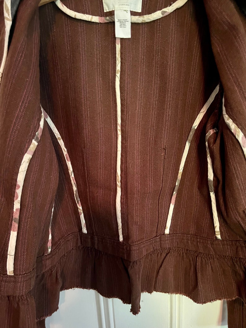 May include: Brown striped jacket with a white floral trim. The jacket has a ruffled hem and a zipper closure.
