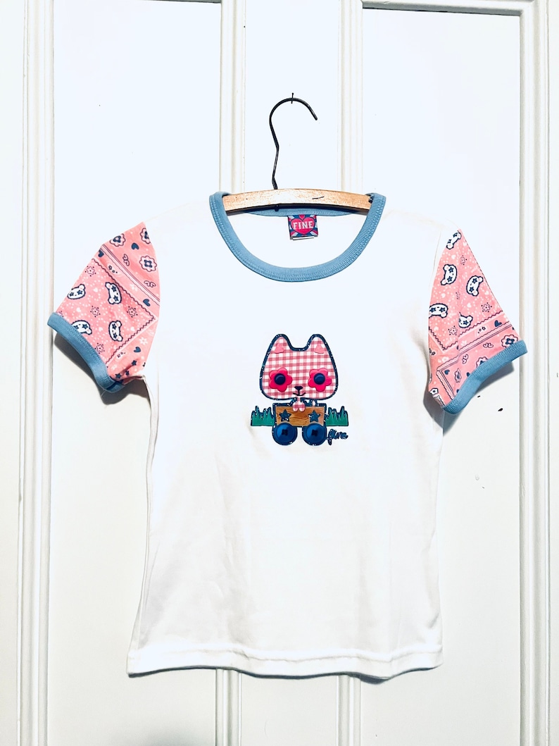 May include: White t-shirt with a blue and pink patterned trim on the sleeves. The shirt features a cartoon cat wearing a red and white checked dress and holding a box with stars. The text "Fine" is on the tag and on the bottom of the cat.