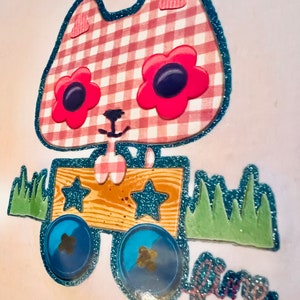 May include: A pink and white gingham cat with blue glitter trim pulls a wooden wagon with blue wheels and stars. The wagon is on a green grass surface.