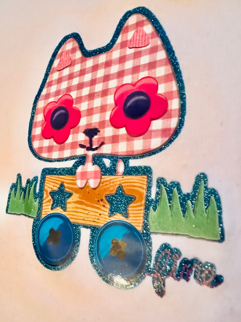 May include: A pink and white gingham cat with blue glitter trim pulls a wooden wagon with blue wheels and stars. The wagon is on a green grass surface.