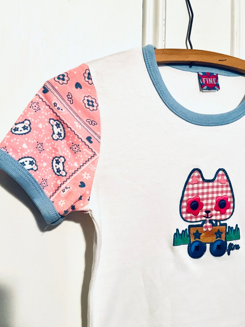 May include: White t-shirt with a blue and pink trim. The sleeves are pink with a bandana print. The front of the shirt features an applique of a cartoon cat wearing a pink and white gingham dress and pulling a wooden wagon. The wagon has two wheels and a blue star on the side. The text "FINE" is on a red tag attached to the shirt.
